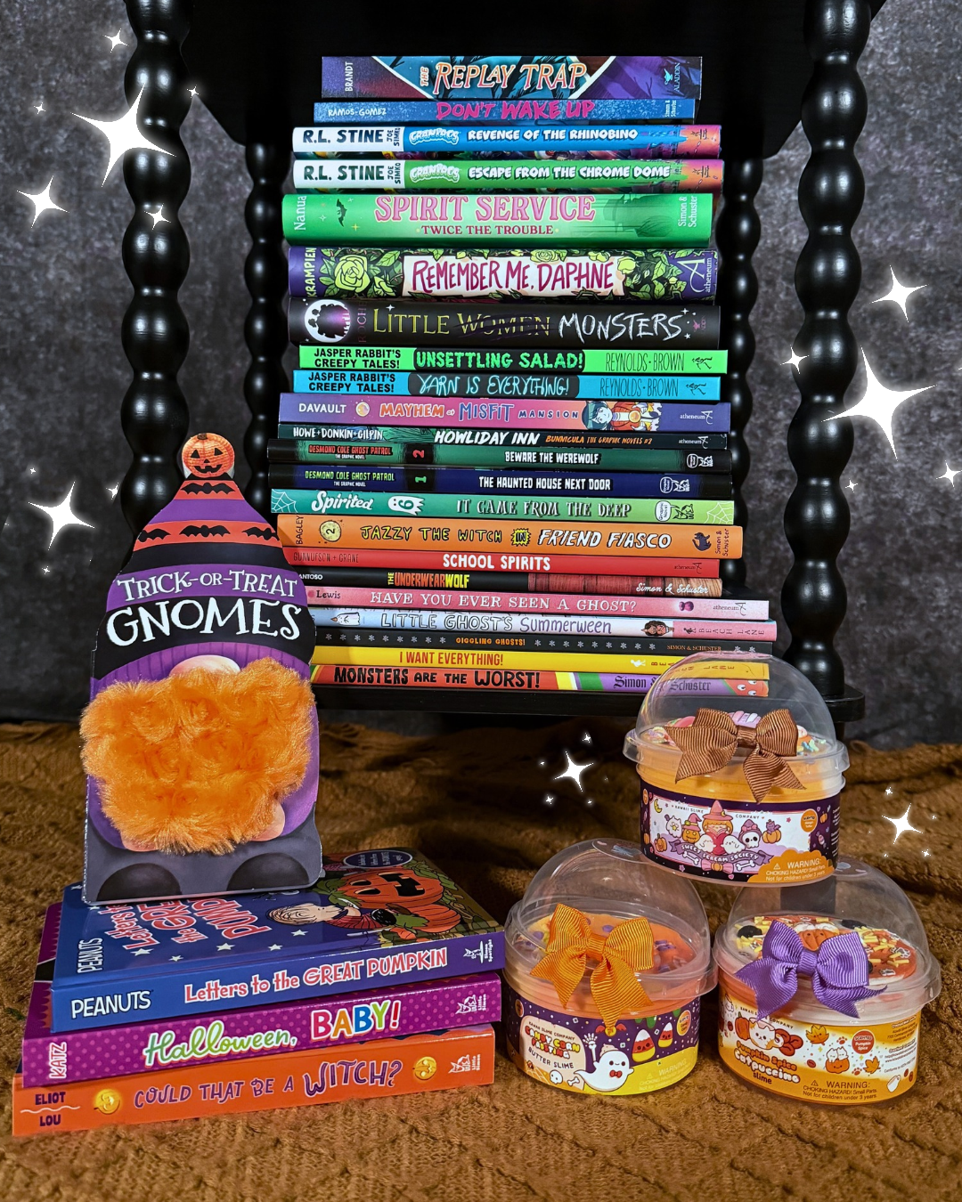 A collection of Halloween-themed books for kids of all ages on a woven orange rug and a black shelf beside three Kawaii Slime Company slime containers.