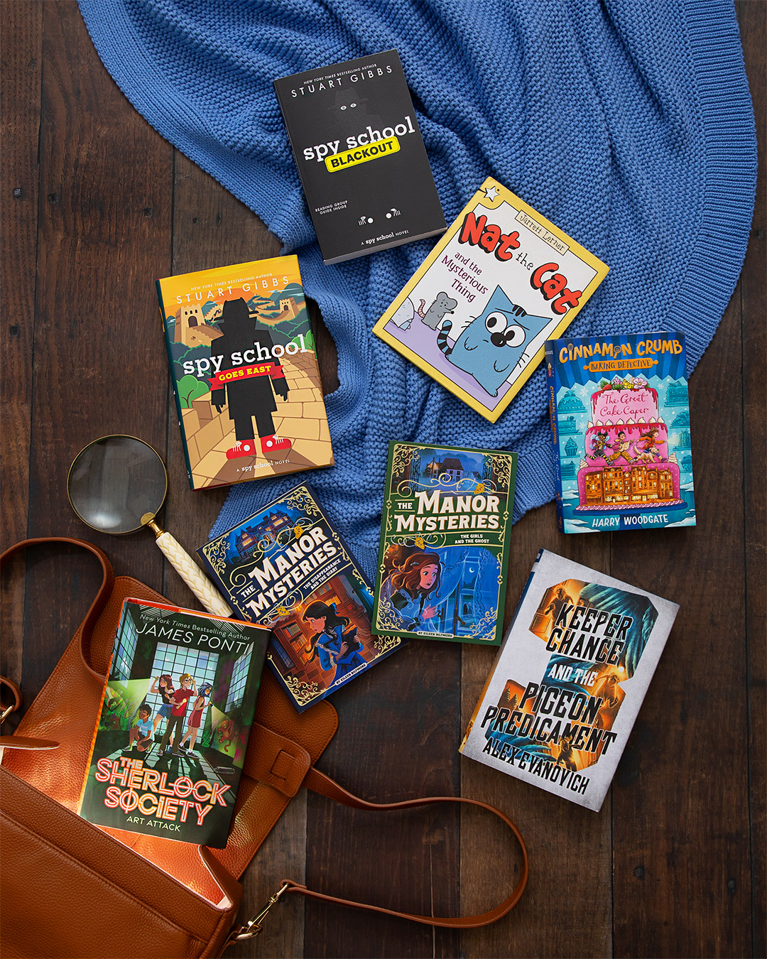 Several middle grade books including SPY SCHOOL GOES EAST and THE GREAT CAKE CAPER spilling out of a brown leather bag, the inside illuminated, onto a blue blanket against a dark wood floor