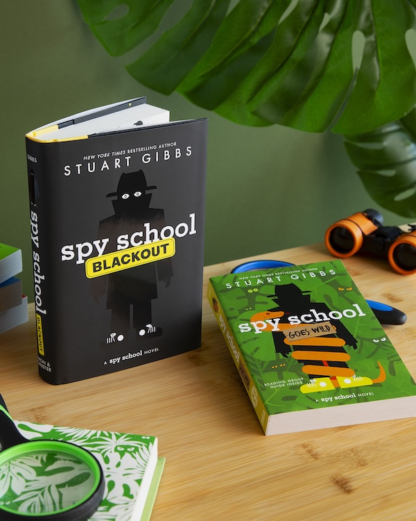 Two Spy School books on a wooden table beside a pair of binoculars and a magnifying glass.