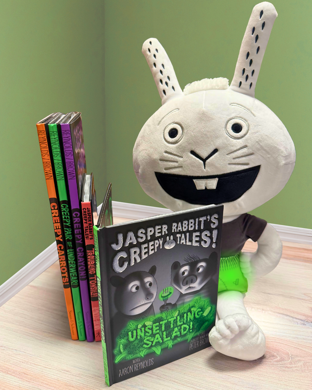 A Jasper Rabbit plush reading UNSETTLING SALAD! by a stack of children's books featuring Jasper Rabbit by Aaron Reynolds and Peter Brown